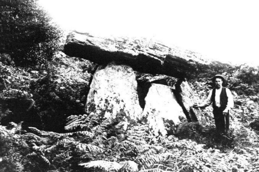 Black & white photo of a man beside a large stone dolmen in a ferned area