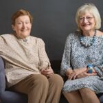 Two women sitting, one (with short brown hair) in a beige jumper and brown pants, and the other (with longer blonde hair and wearing glasses) in a blue floral dress