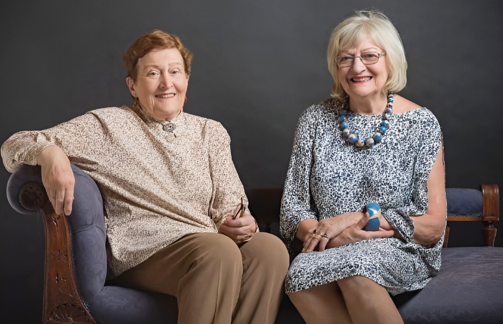 Two women sitting, one (with short brown hair) in a beige jumper and brown pants, and the other (with longer blonde hair and wearing glasses) in a blue floral dress