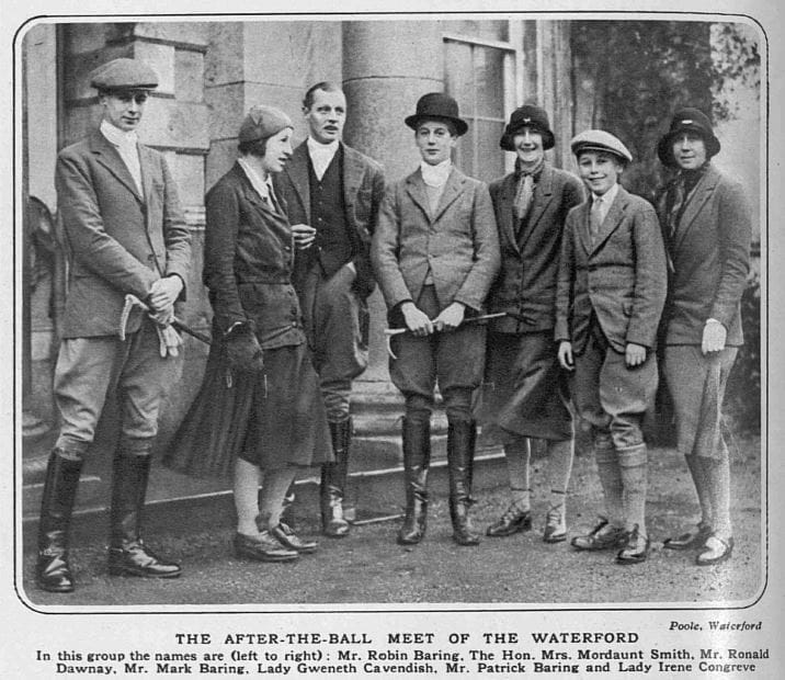 Black & white photograph of people outside a large house, most in hunting attire.