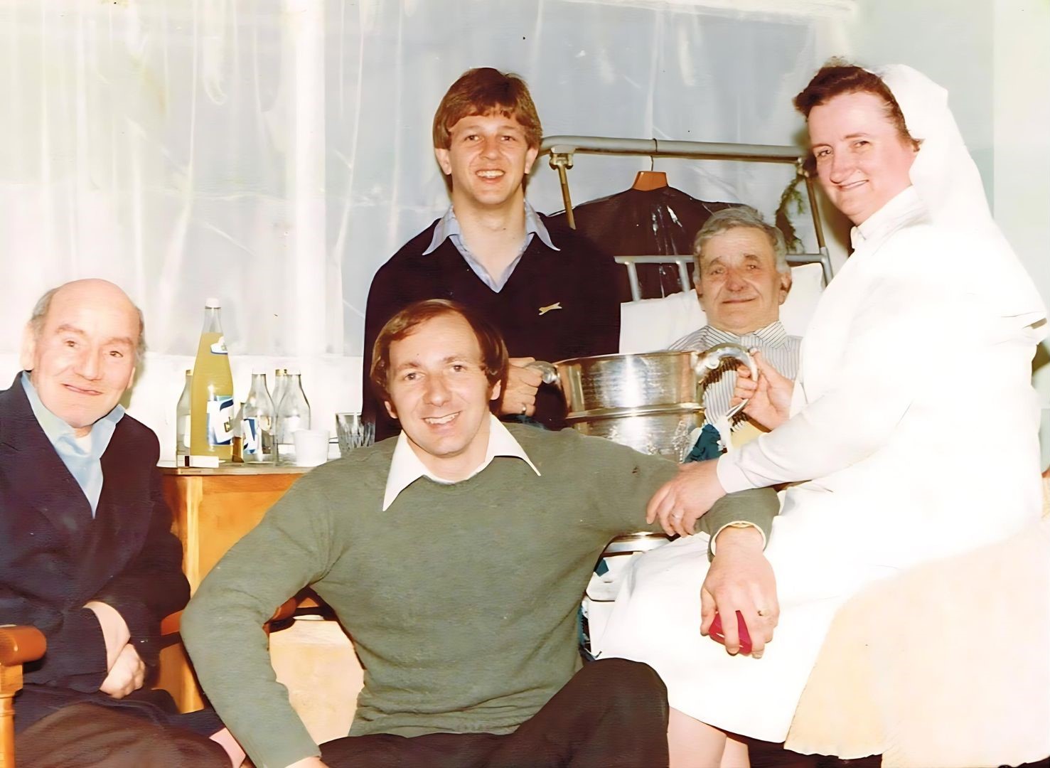 Colour photograph of two men and a female nurse at the bedsides of two elderly hospital patients, with everyone smiling.