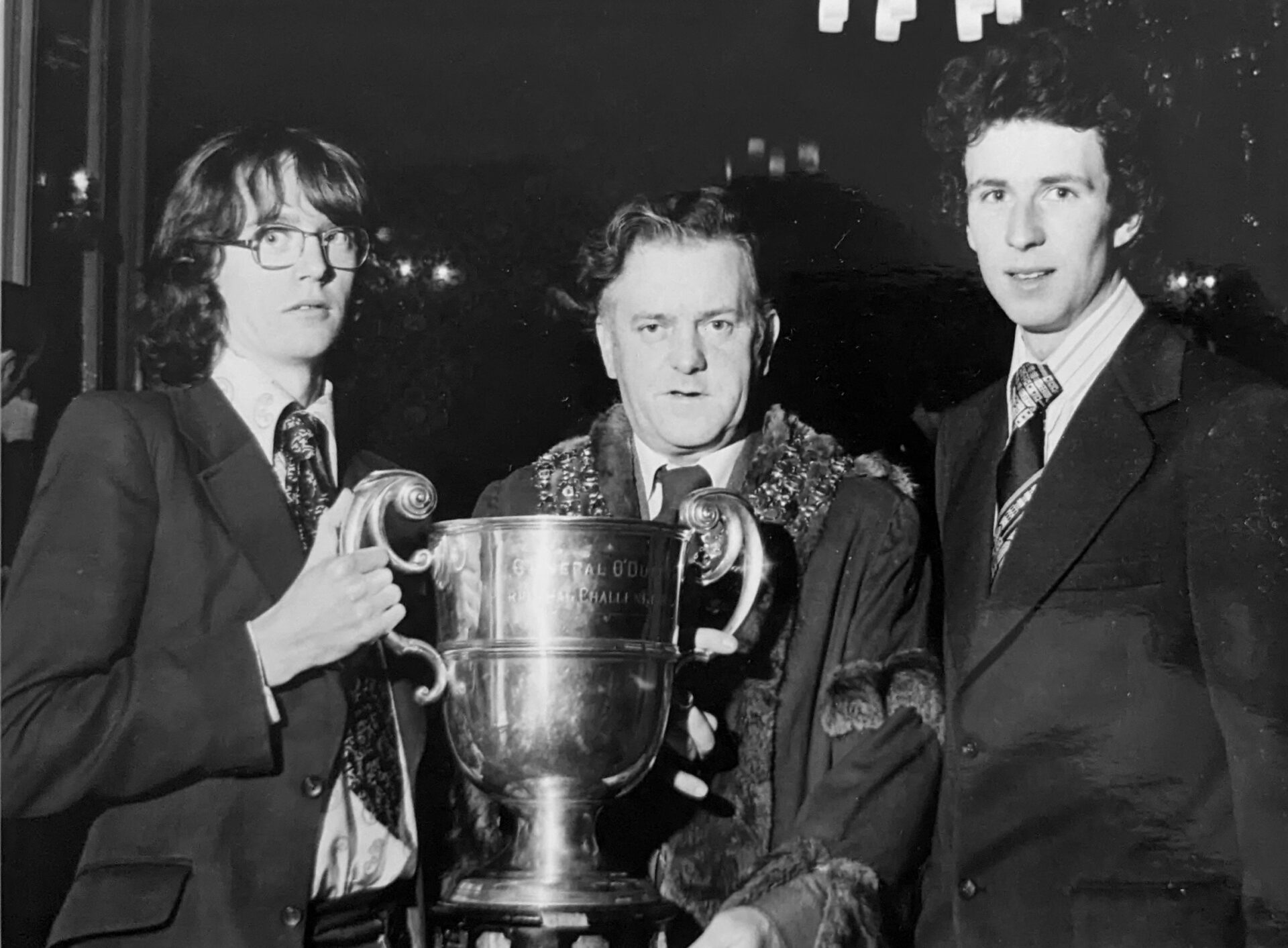 Black & white photo of a man wearing the robes of office and mayoral chain with two men wearing suits and one (wearing glasses) is holding a large cup