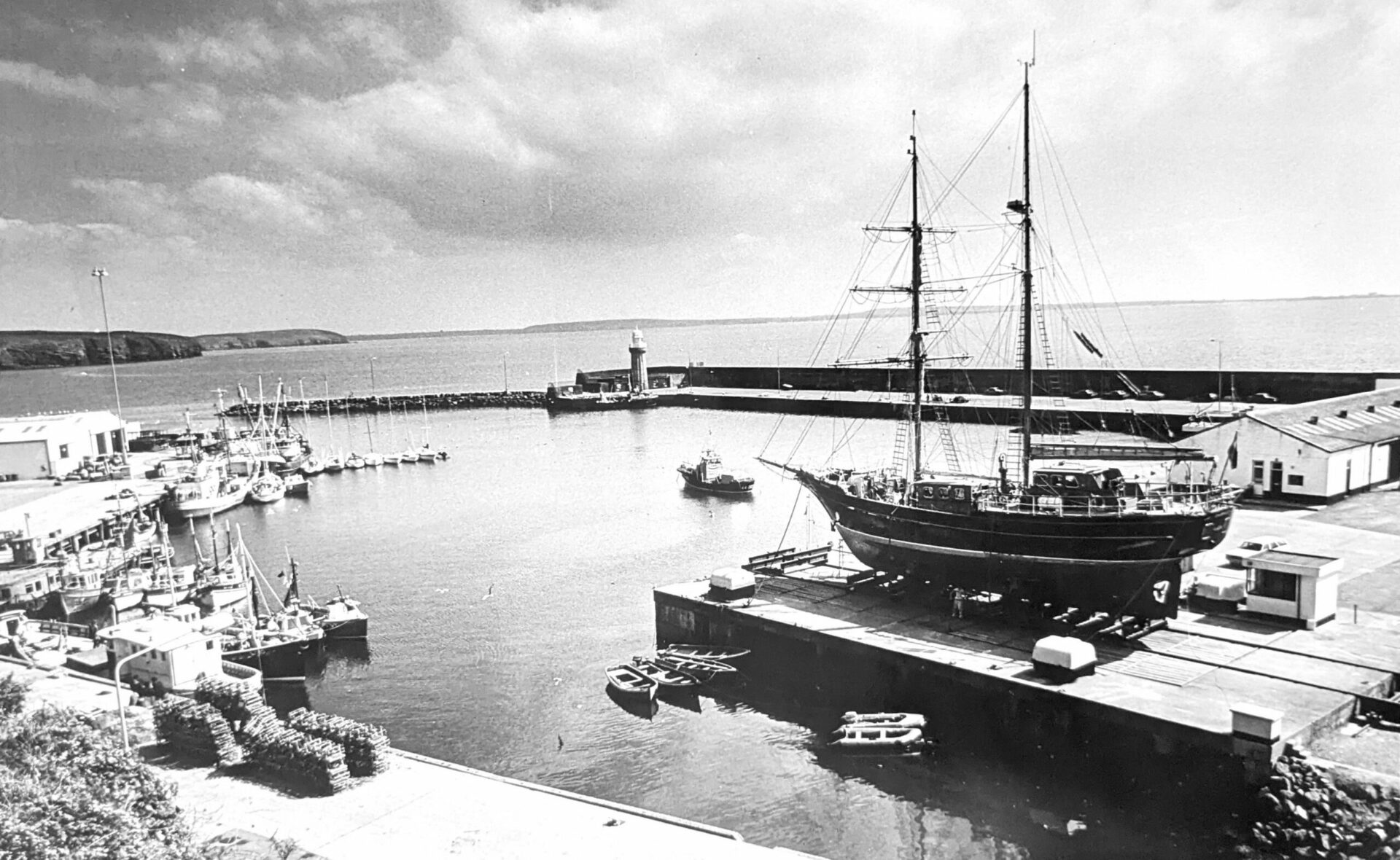 Black & white photograph of an old wooden sailing ship with its sails down in dock at a fishing port with other boats also in port and a lighthouse on the pier