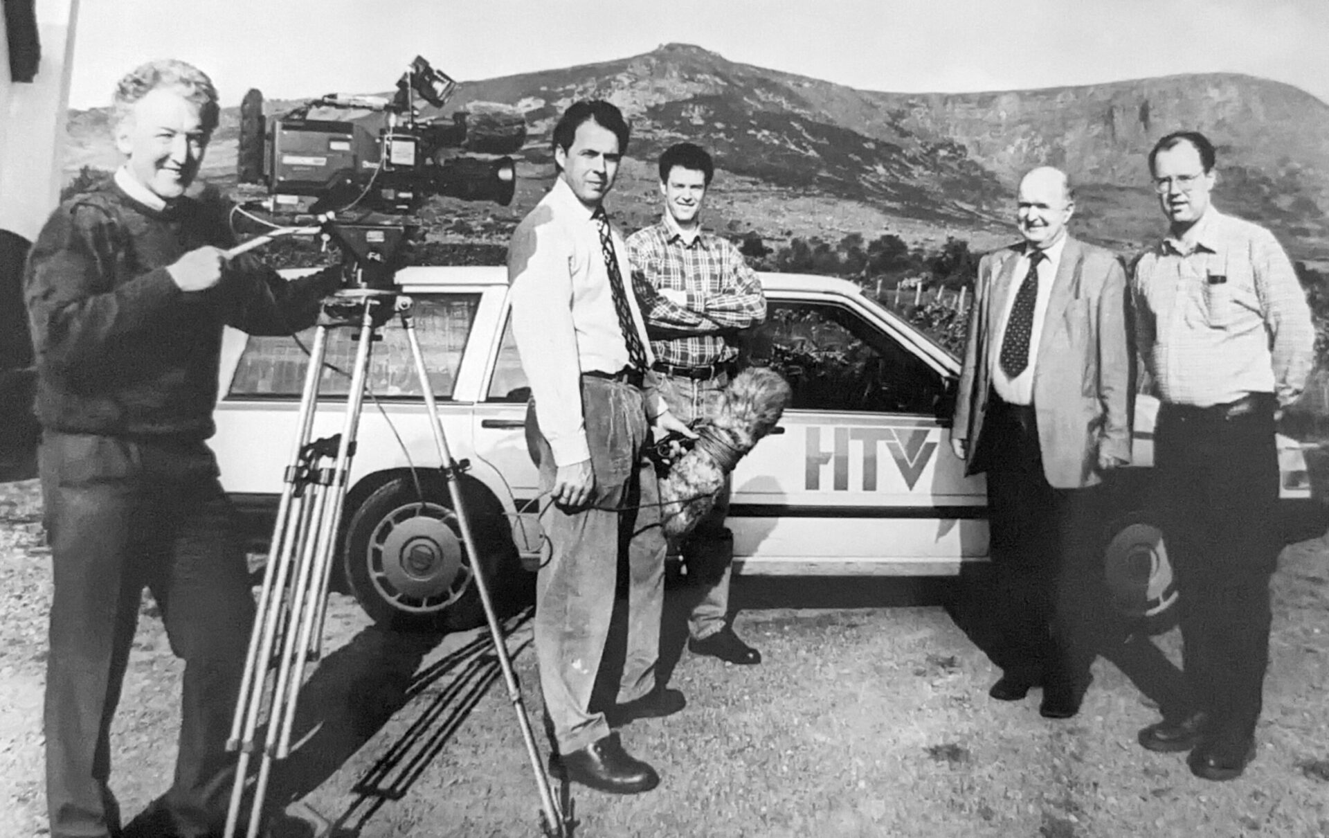 Five men, three of them with TV camera and audio recording equipment, beside a light-coloured stationwagon car bearing the logo "HTV" on the door, with a mountain range in the background