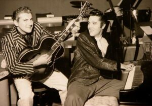 A black & white photo of the musical entertainers Liberace (holding a guitar) and Elvis (on the piano) in 1956