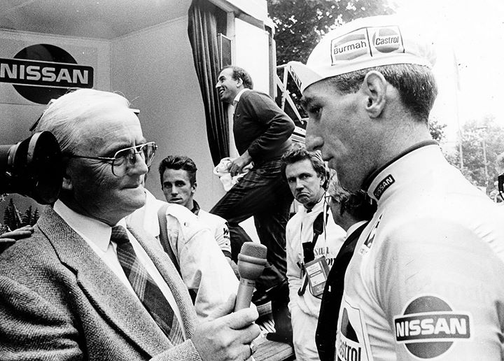 Black & white photo of a white-haired, bespectacled journalist interviewing a cap-wearing cyclist after a race with other riders in the background